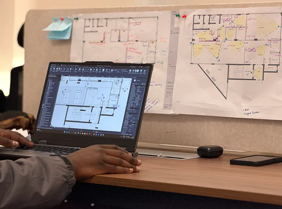 A laptop running CAD beside hand marked floor plans pinned to a desk screen